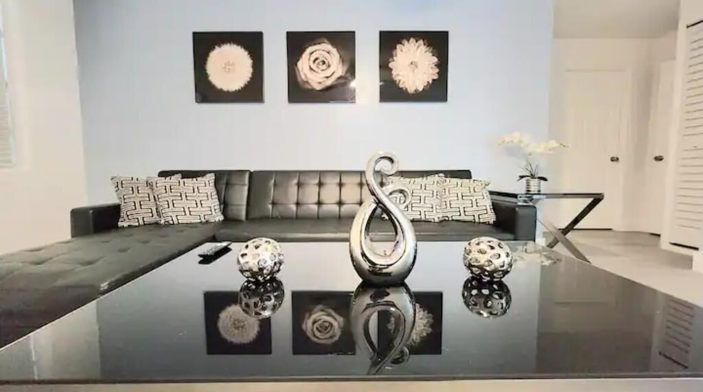 A modern living room with a glass coffee table, a grey sofa with patterned cushions, and three wall-mounted decorative pieces.