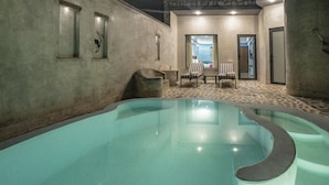 Une piscine intérieure avec un sol carrelé, un espace détente avec des fauteuils et une table, et une porte donnant sur une autre pièce.