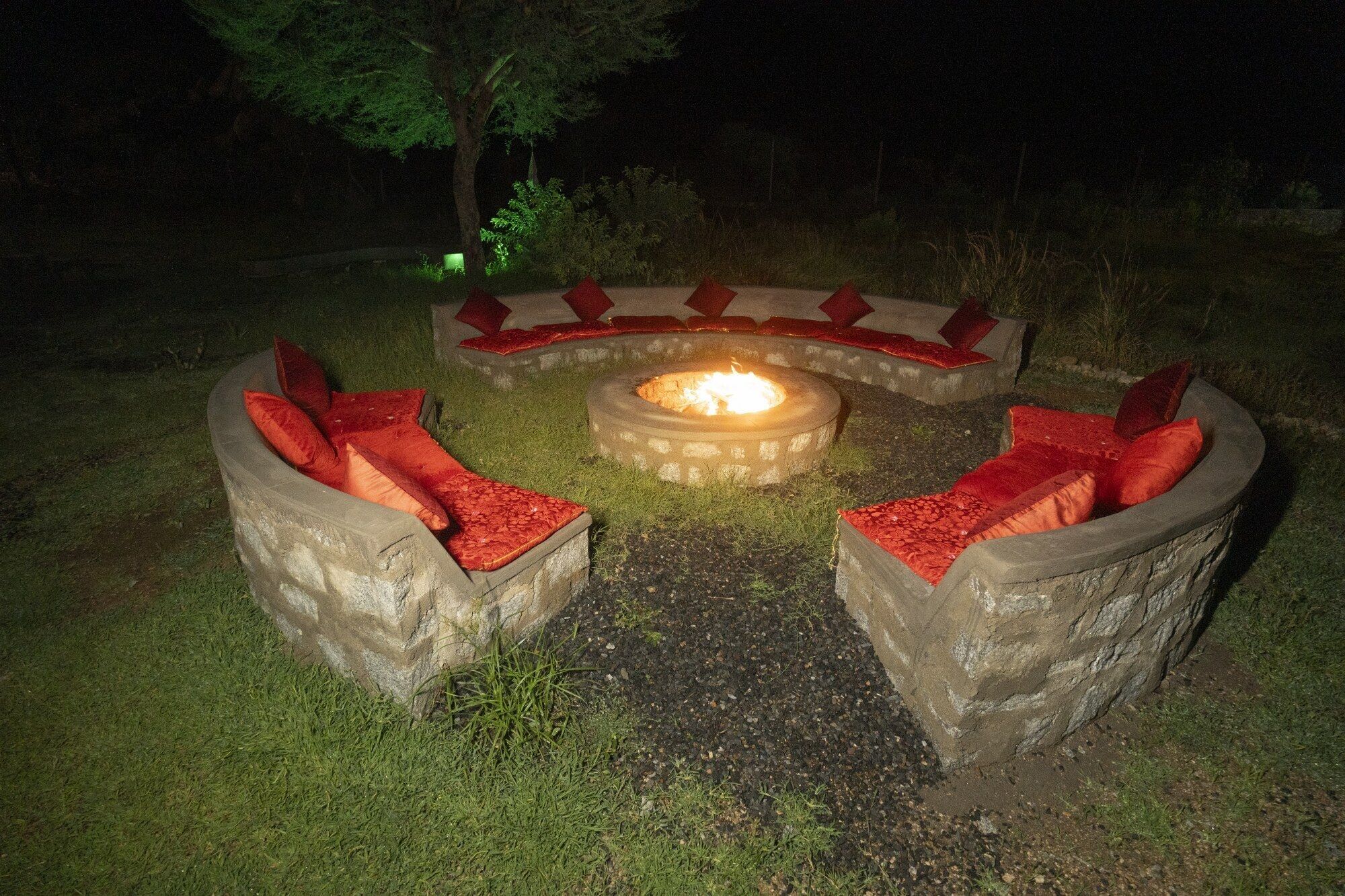 Hoguera de piedra circular con cojines rojos y una llama central, rodeada de un césped de noche.
