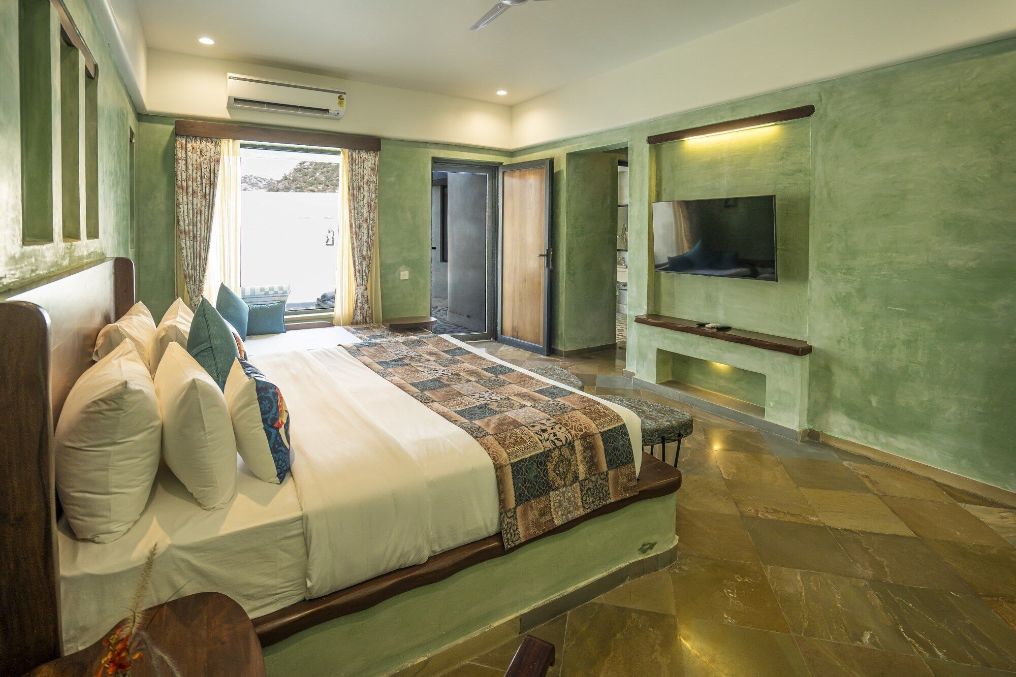 A spacious bedroom with a large bed, a TV mounted on the wall, and a view of the outdoors through the door.