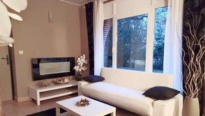 Uma sala de estar moderna com um sofá branco, uma mesa de centro, uma TV de tela plana sobre um suporte branco e uma janela com vista para árvores.