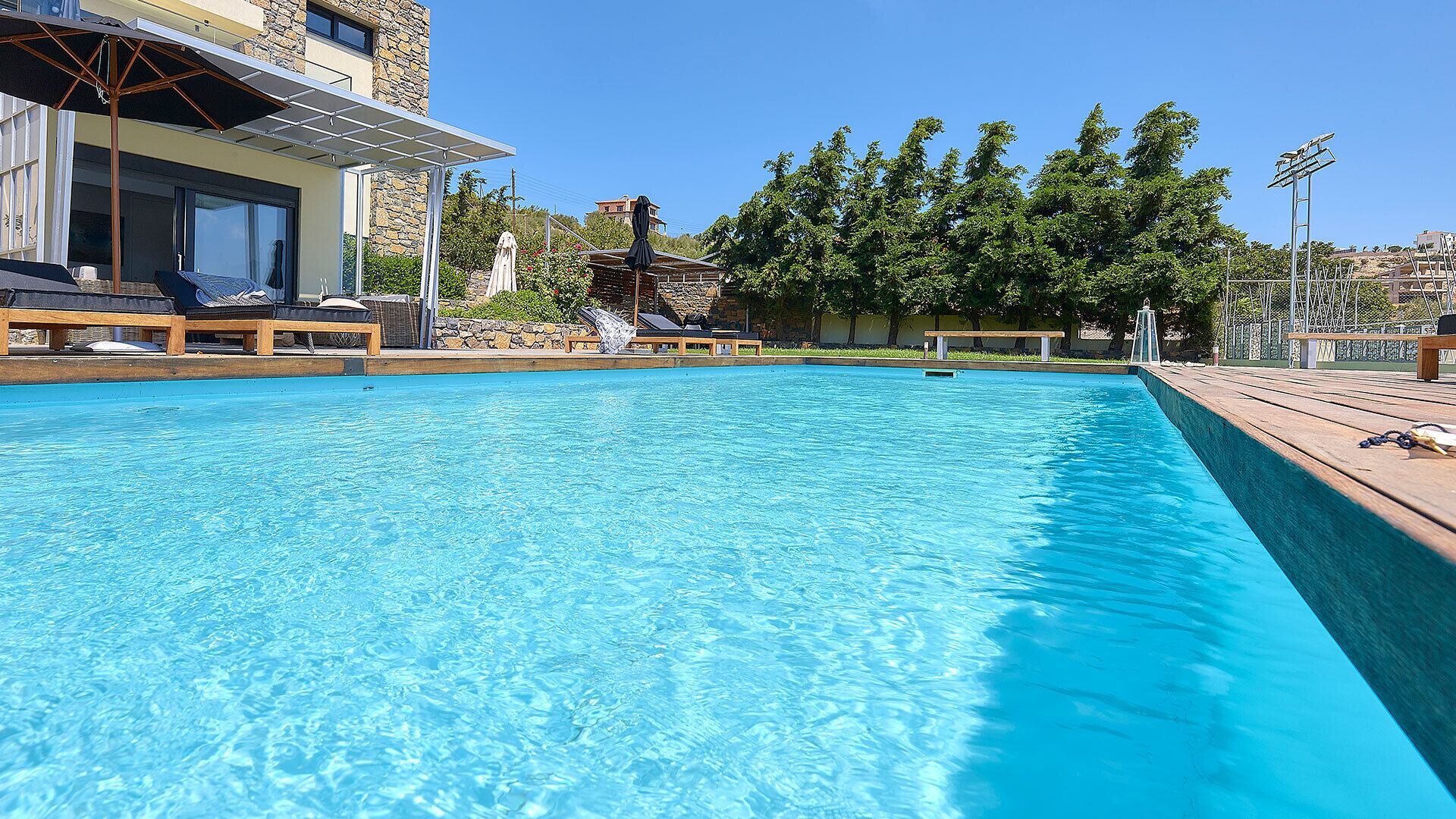 A clear blue pool with wooden deck chairs and umbrellas, surrounded by a stone building and trees.