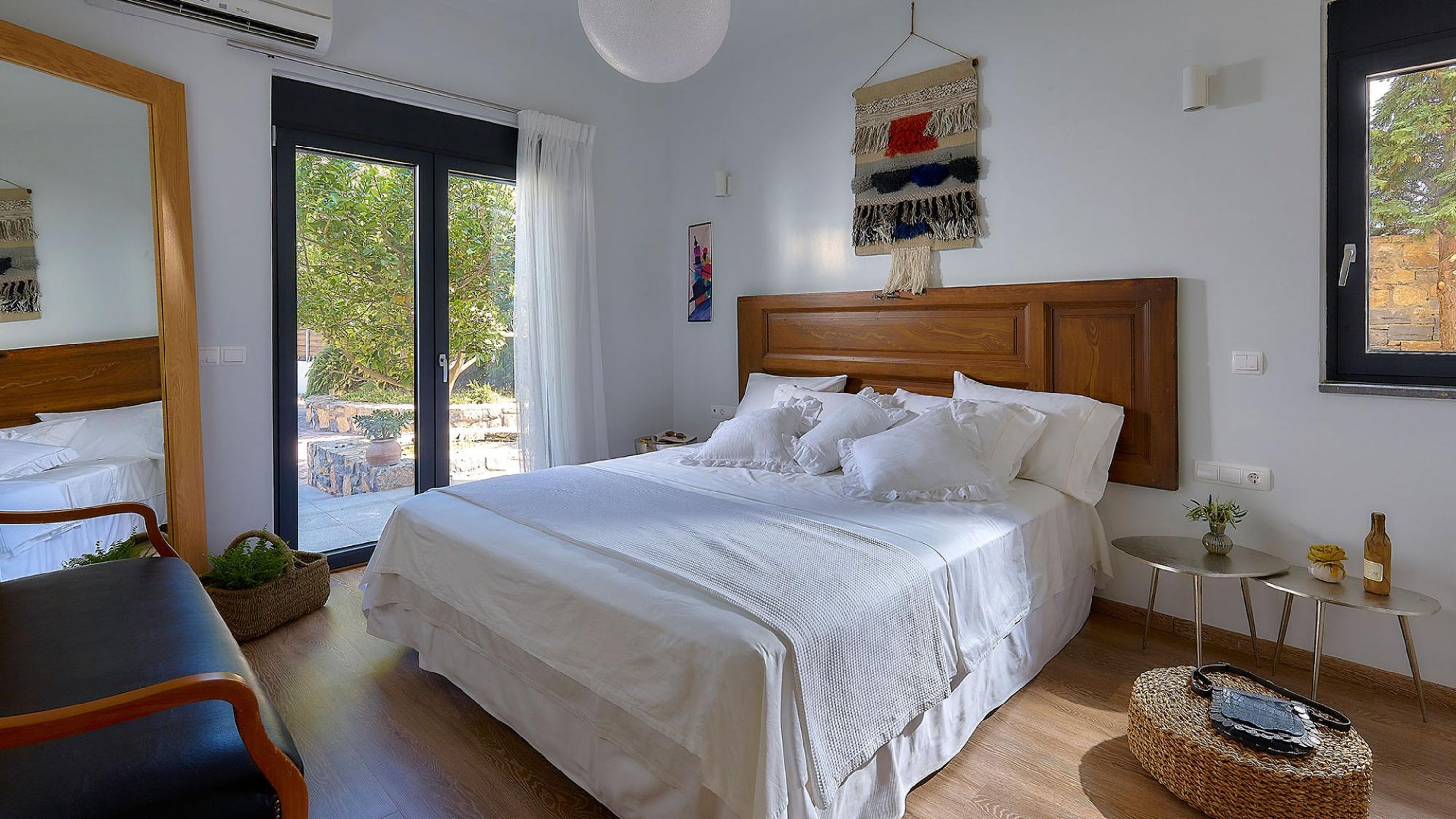 A bedroom with a large bed, a small table, a chair, and a view of the outdoors through a sliding door.