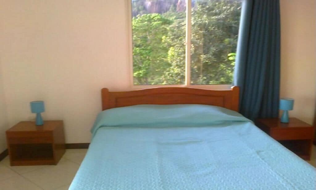 Ein Schlafzimmer mit einem Bett, zwei Nachttischen, einem Fenster mit Blick auf Bäume und einem Vorhang.