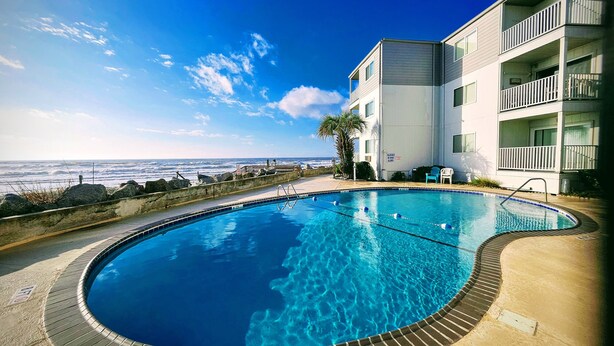 A swimming pool with a view of the ocean, a building with balconies, and a clear sky. - OCEAN FRONT @ CHERRY GROVE POINT - SALT AIR, SEA BREEZES, GORGEOUS SUNRISES (North Myrtle Beach)