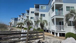 A row of multi-story residential buildings with balconies, a wooden fence, and palm trees. - OCEAN FRONT @ CHERRY GROVE POINT - SALT AIR, SEA BREEZES, GORGEOUS SUNRISES (North Myrtle Beach)