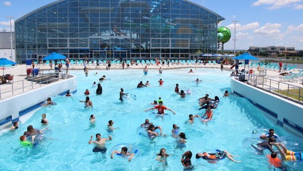 A large indoor swimming pool with people swimming and using inflatable rings, surrounded by a modern building with large glass windows. - 4 BR/2 BA walking distance to AT&T, Ranger Stadium, Texas Live, Six Flags (Arlington)