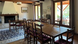 A dining area with a large wooden table, several chairs, a fireplace, and a view of the outdoors through large windows. - Les Costes, Calvignac. Large house with pool. Close to the river. (Calvignac)