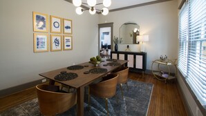 Un coin repas avec une longue table en bois, six chaises, un lustre, des œuvres d’art encadrées au mur et une fenêtre avec des stores.
