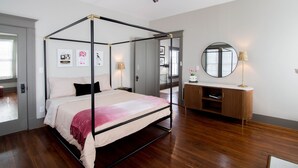 Un lit à baldaquin avec un cadre en bois, une table de chevet avec une lampe, un miroir rond accroché au mur et une armoire en bois avec un plan de travail blanc.