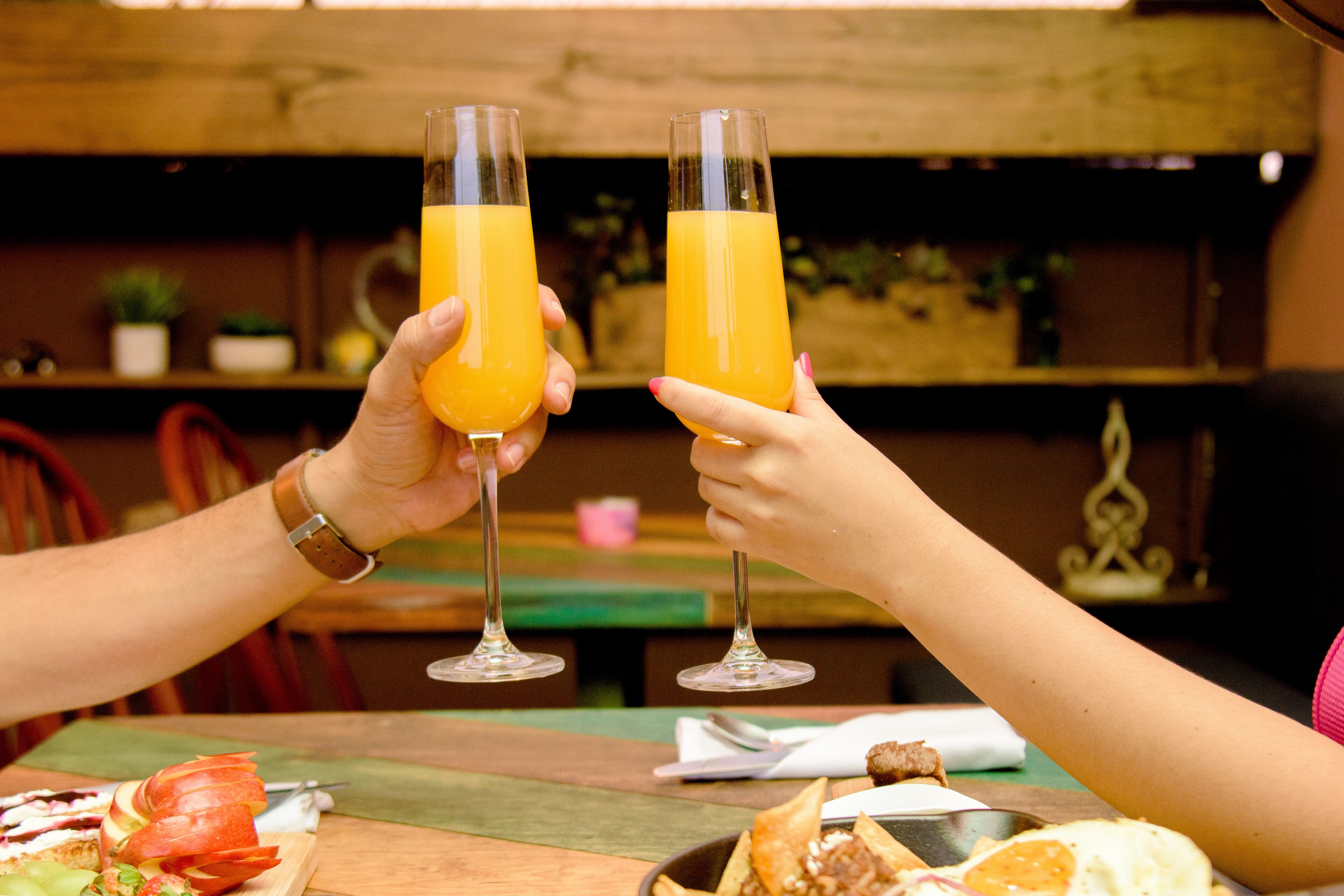 Two people holding glasses of orange juice at a wooden table with food items and a spoon.