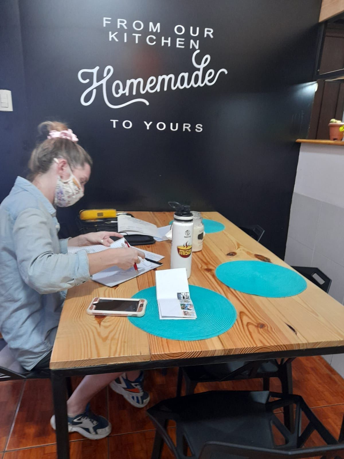 Una persona seduta a un tavolo di legno con un quaderno, uno smartphone e una tazza di caffè. Sullo sfondo, un cartello con su scritto “DALLA NOSTRA CUCINA Fatto in casa PER VOI.”