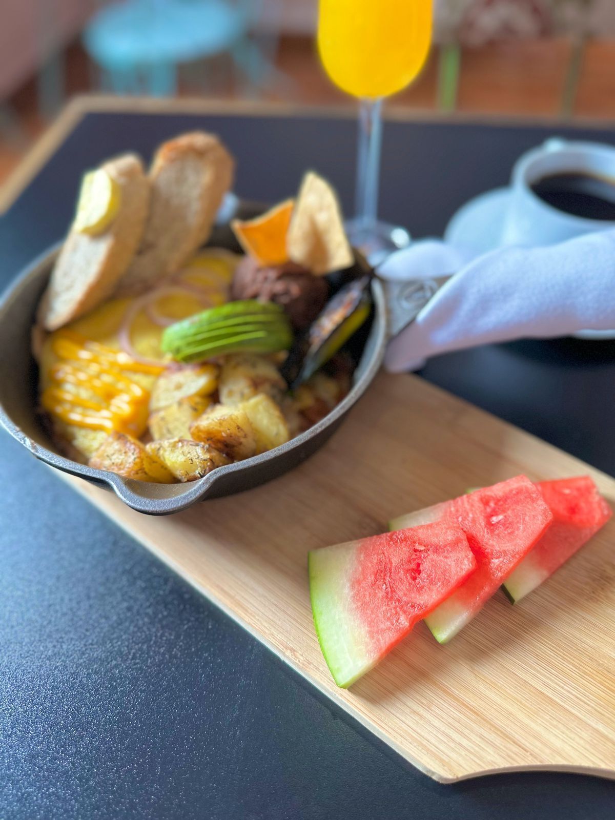 A breakfast dish in a cast iron skillet, accompanied by sliced watermelon and toast on a wooden board.