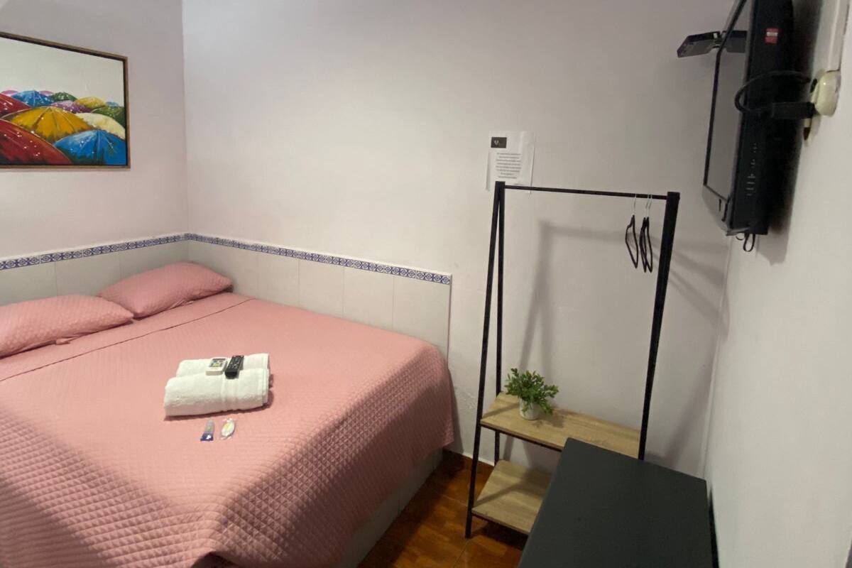 A bedroom with a bed, a bedside table, a hanging lamp, a painting, and a black suitcase on the wall.