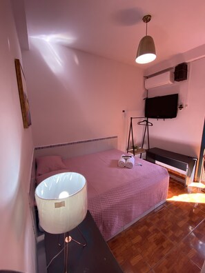 A bedroom with a bed, a bedside table with a lamp, a TV on the wall, and a wooden floor.