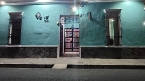 A building facade with a teal exterior, a central door with a sign, and two windows with metal bars. - HOSTAL VELVET  (Santa Ana)