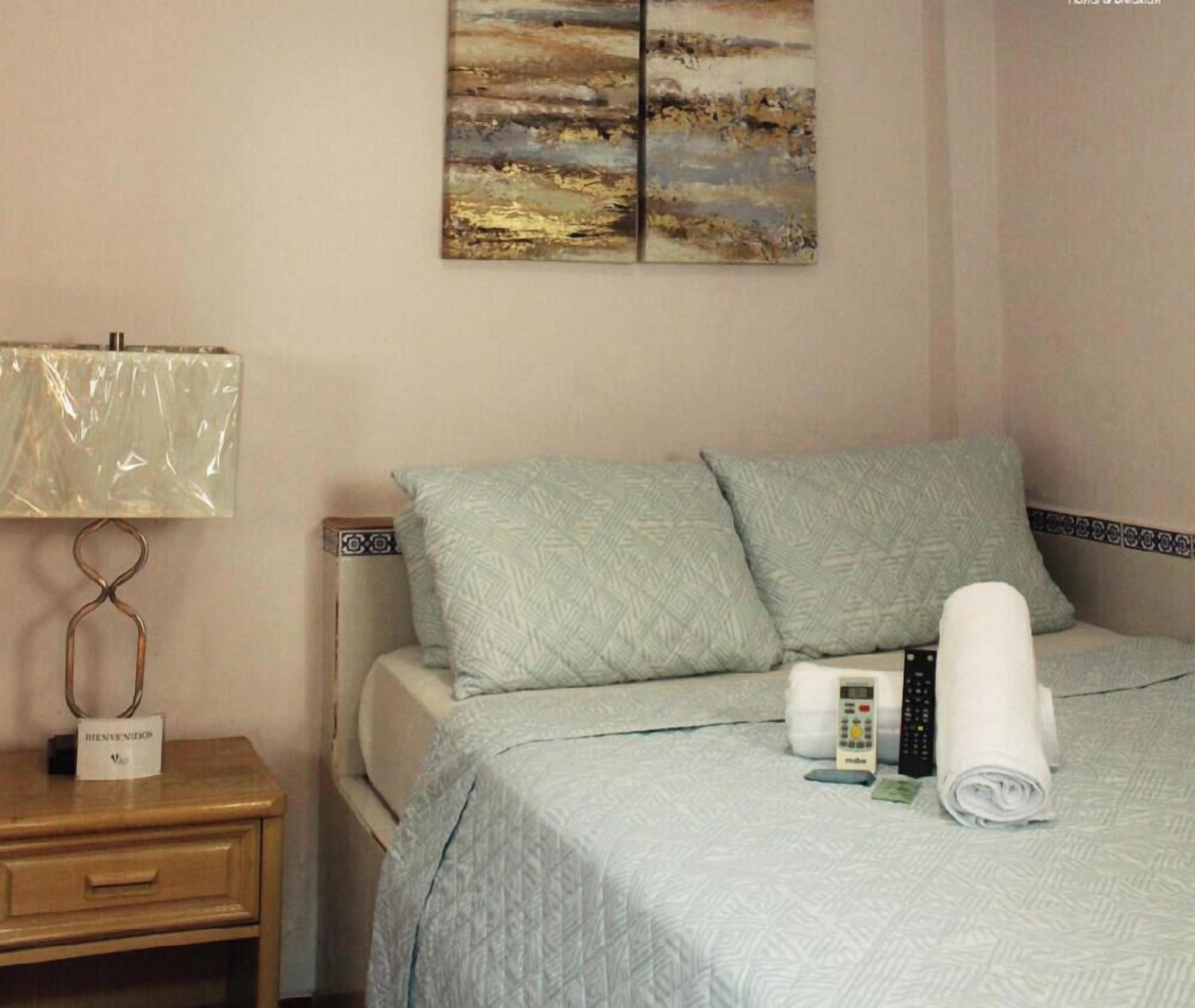 A hotel room with a bed, a nightstand, a lamp, a remote control, and a towel.