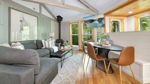 Ein gemütliches Wohnzimmer mit einer Couch, einem Esstisch mit Stühlen, einem Kamin und großen Fenstern mit Blick ins Grüne.