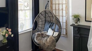 A room with a white chandelier, a round wicker chair with cushions, a large window with blinds, and a decorative mirror.