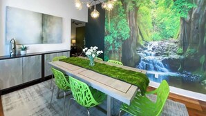 A dining area with a long table, green chairs, a chandelier, and a large wall mural of a forest waterfall.