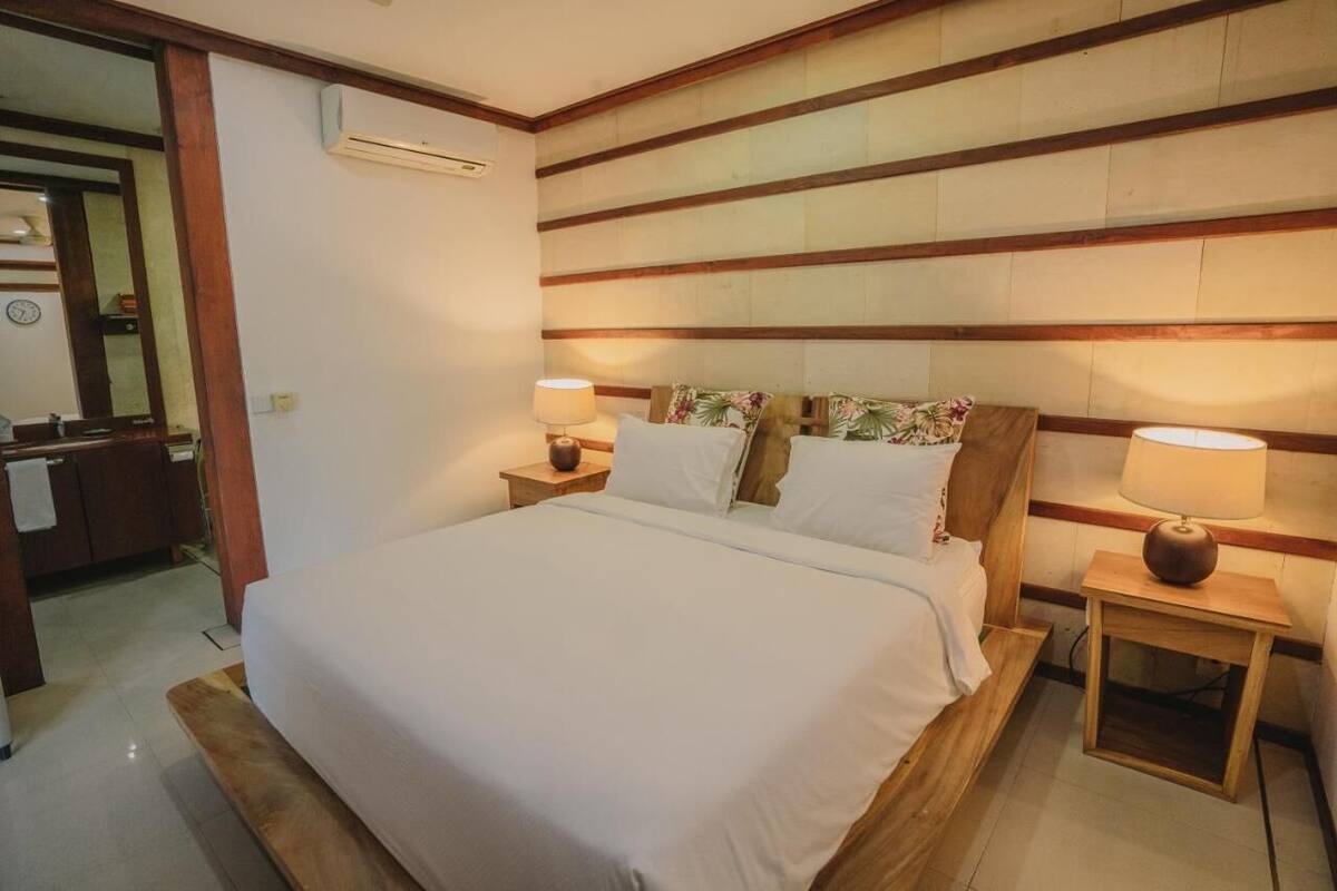 A hotel room with a large bed, wooden headboard, two bedside tables with lamps, and a small bathroom visible in the background.