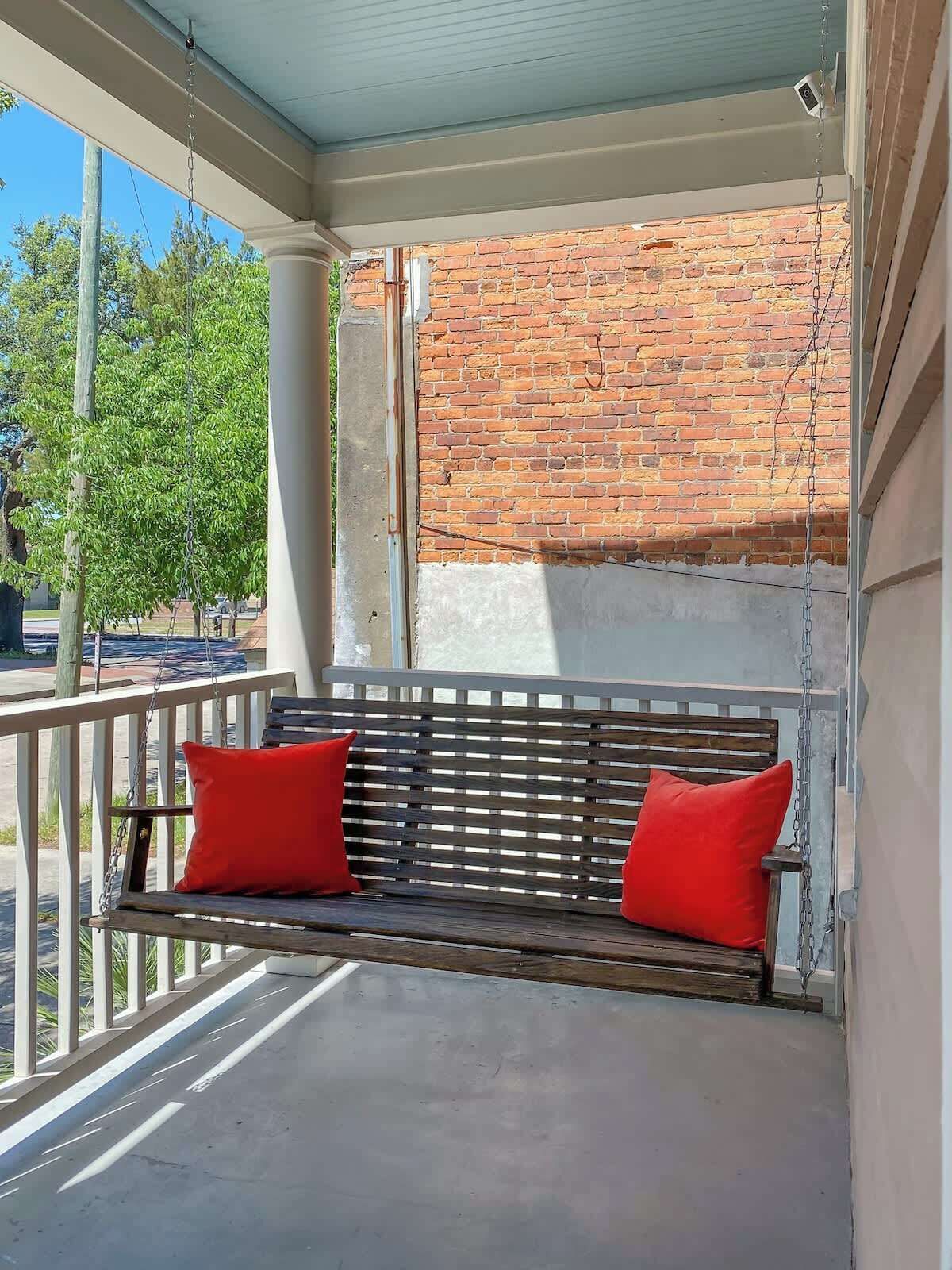 A porch with a wooden bench and red cushions, a brick wall in the background, and a clear view of the outdoors.