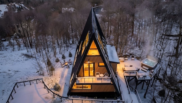 Een moderne cabin met een puntig dak, omringd door met sneeuw bedekte grond en bomen, met een gezellig interieur met zitplaatsen en een hottub.