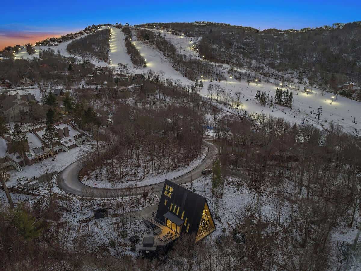 Une station de ski enneigée, avec une route sinueuse, un bâtiment noir à l’architecture unique, doté de fenêtres triangulaires, et un télésiège en arrière-plan.