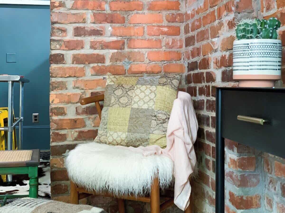 A cozy corner with a wooden chair, a patterned cushion, and a small potted plant on a brick wall.