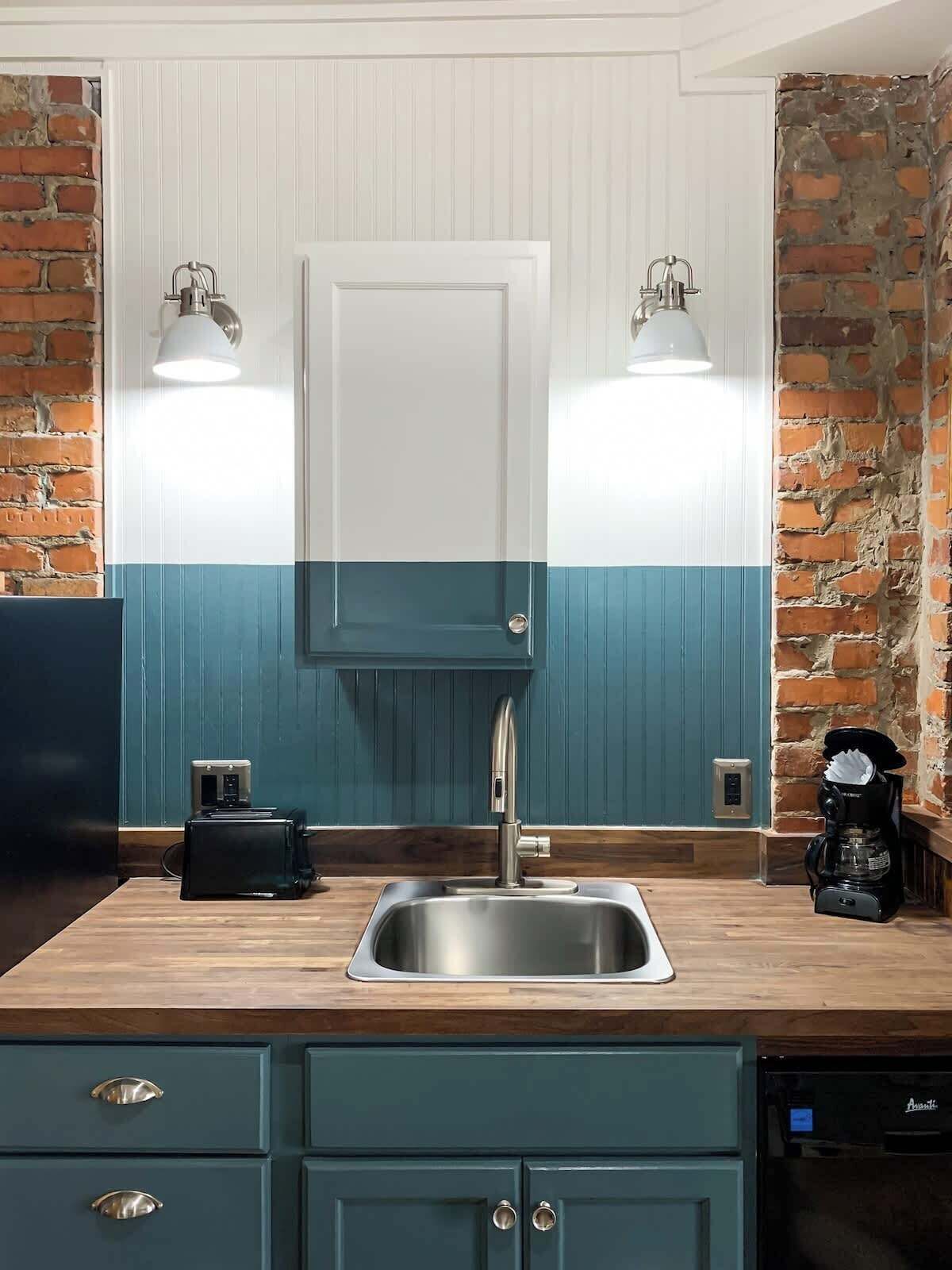 A kitchen with a stainless steel sink, wooden countertop, teal cabinetry, and a brick wall.