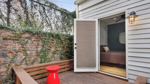 Une terrasse en bois avec un tabouret rouge, un banc et une porte entrouverte donnant sur une pièce où se trouvent un lit et un luminaire fixé au mur.