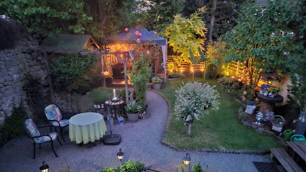 Um pátio com jardim, um gazebo, área de estar e iluminação decorativa.