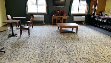 A room with a patterned carpet, a wooden table, a TV mounted on the wall, and a kitchenette area.