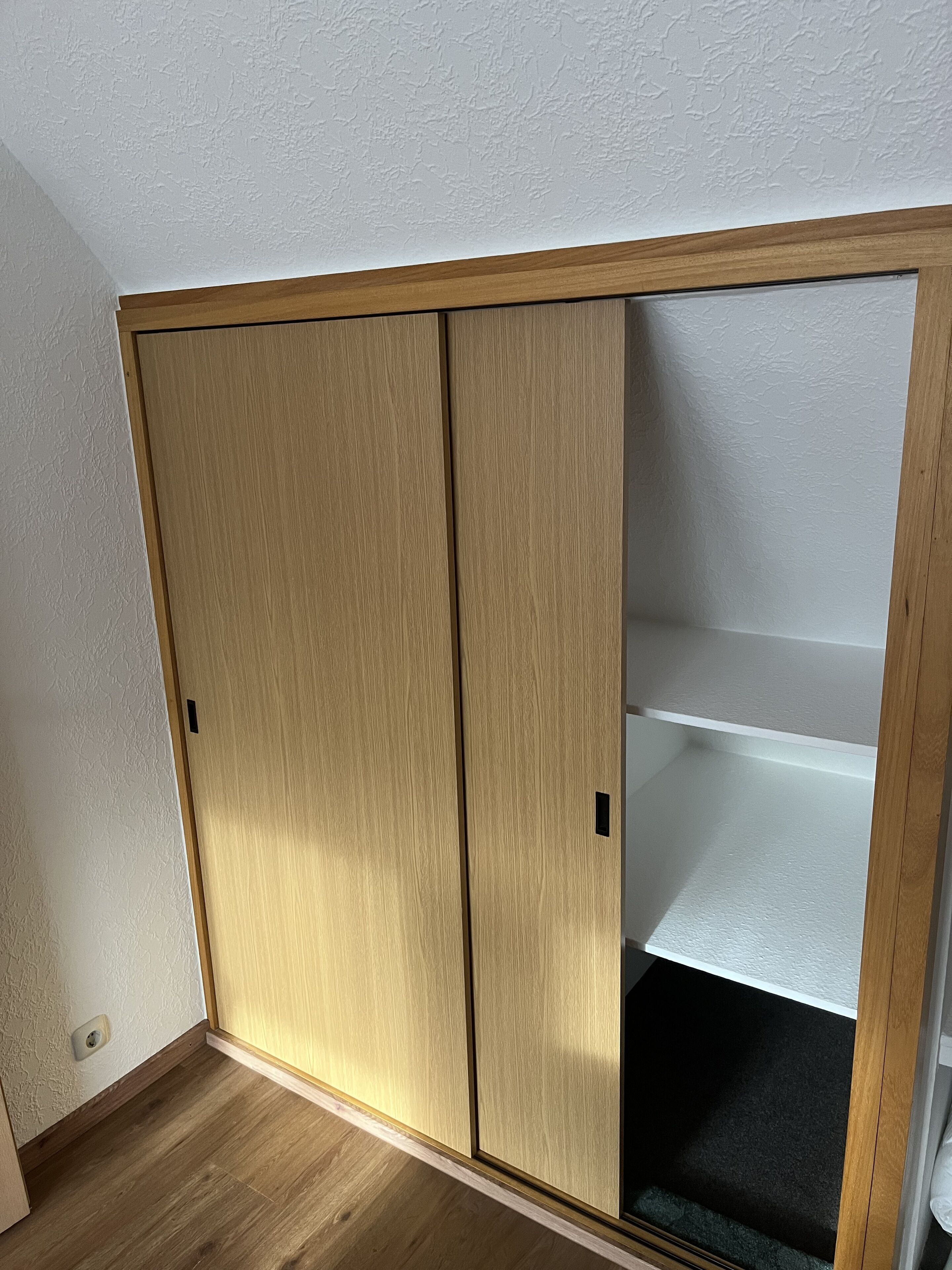 A room with a wooden wardrobe, a white textured wall, and a wooden floor.