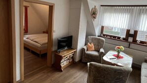 Habitación compacta con cama, televisor, sillón y una mesita con un florero.