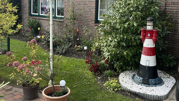 Una casa de ladrillo con una antena parabólica, un jardín con plantas en macetas, un farol blanco y un modelo de faro rojo y blanco.