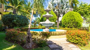 Ein Garten mit einem zentralen Brunnen, umgeben von üppigem Grün und blühenden Pflanzen.