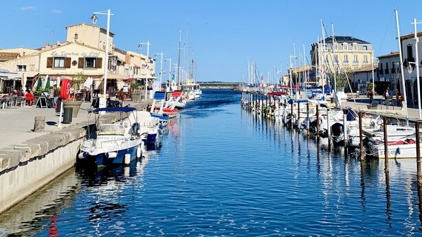 Ein Yachthafen mit vertäuten Booten, ein Gebäude mit der Aufschrift 'La Taverne du Port' auf der linken Seite und einem strahlend blauen Himmel.