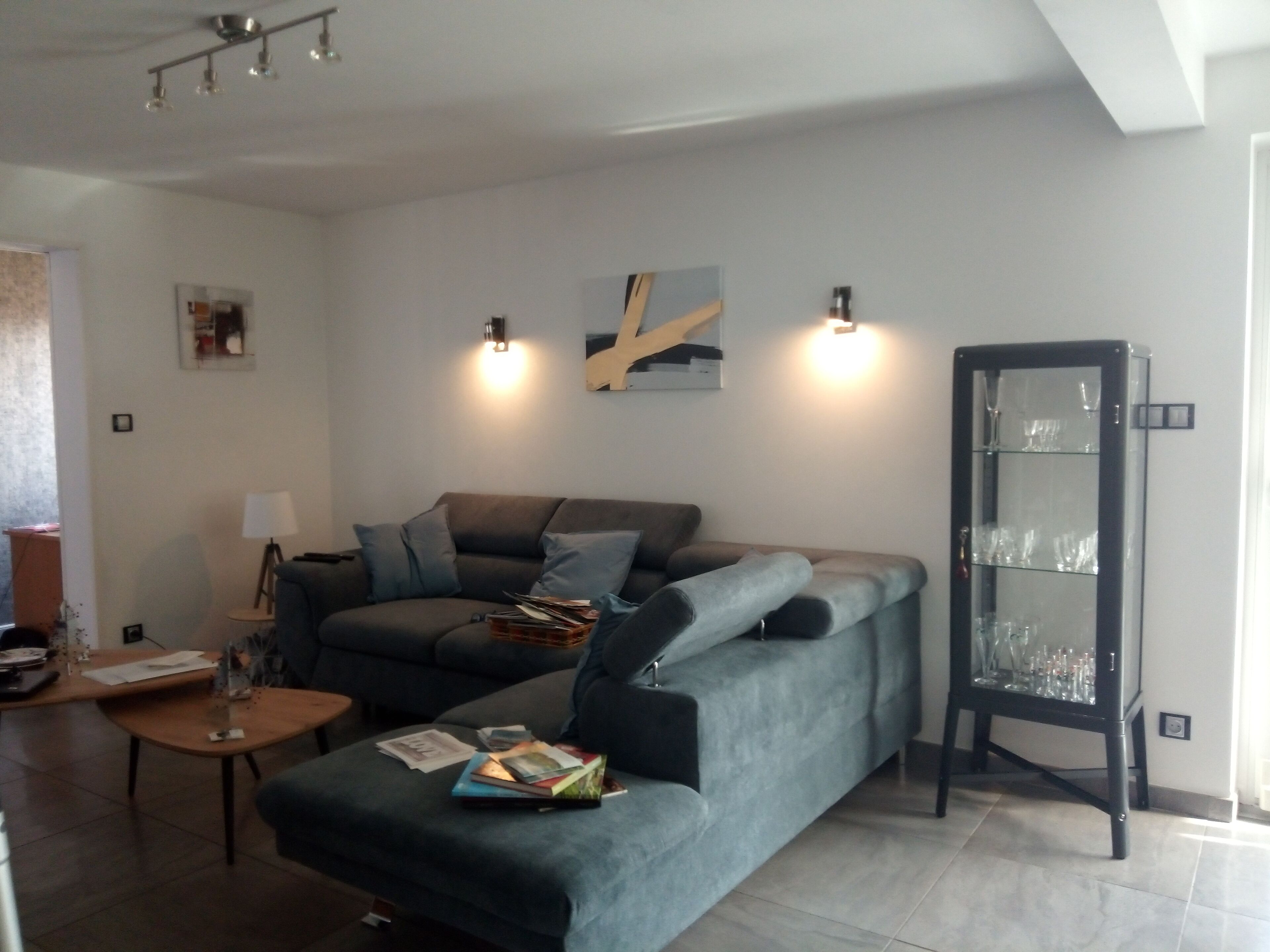 Ein Wohnzimmer mit grauem Sofa, einem kleinen Holztisch und einer Vitrine mit Weingläsern.