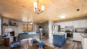 Amplia sala de estar con chimenea, mesa de comedor con cuencos azules y copas de vino, cocina con armarios blancos y electrodomésticos de acero inoxidable, y techo de madera.