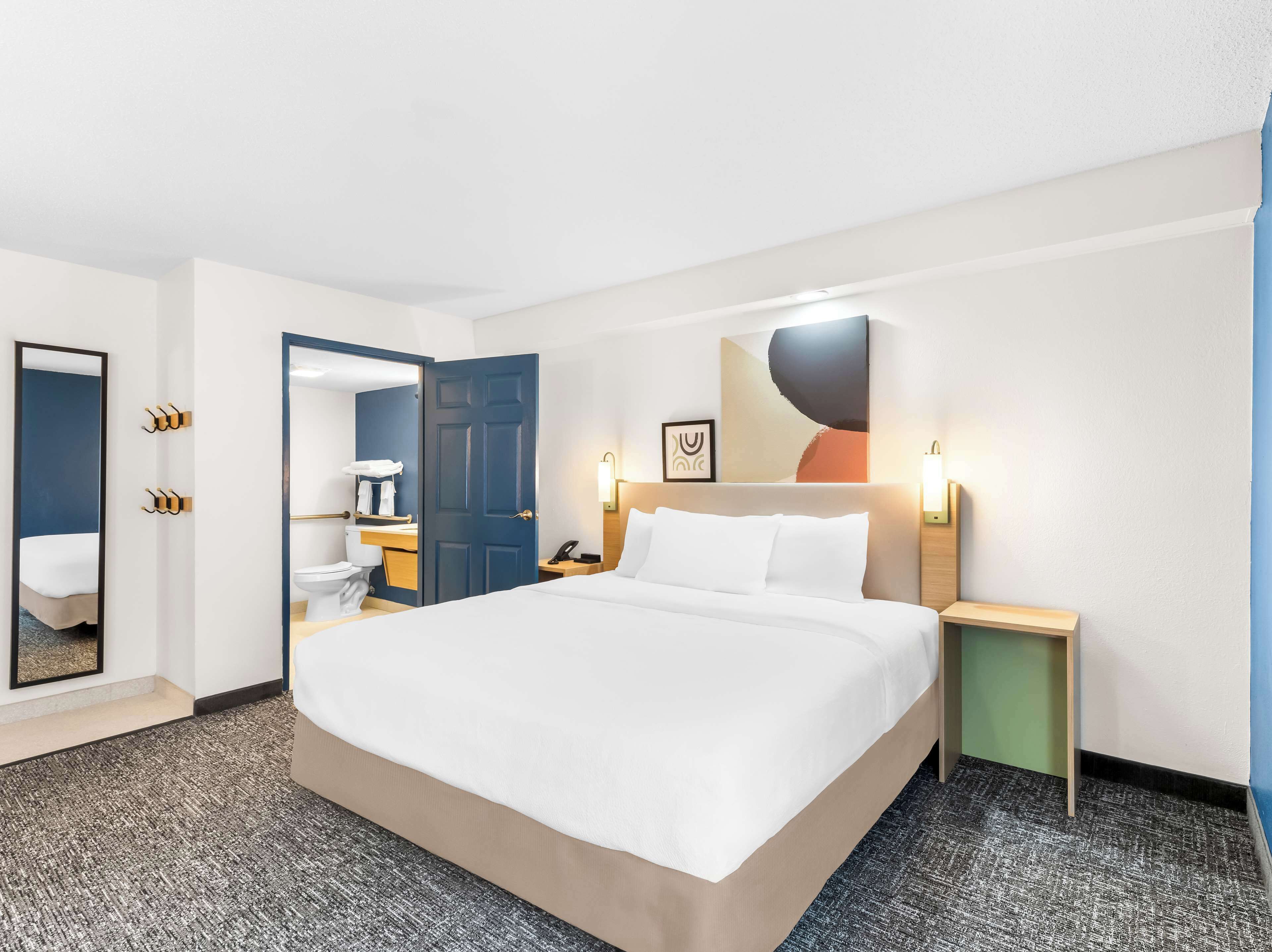 A hotel room with a large bed, a bedside table, a nightstand, a mirror, a bathroom with a toilet and sink, and a wall-mounted artwork.