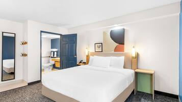 A hotel room with a large bed, a bedside table, a nightstand, a mirror, a bathroom with a toilet and sink, and a wall-mounted artwork.