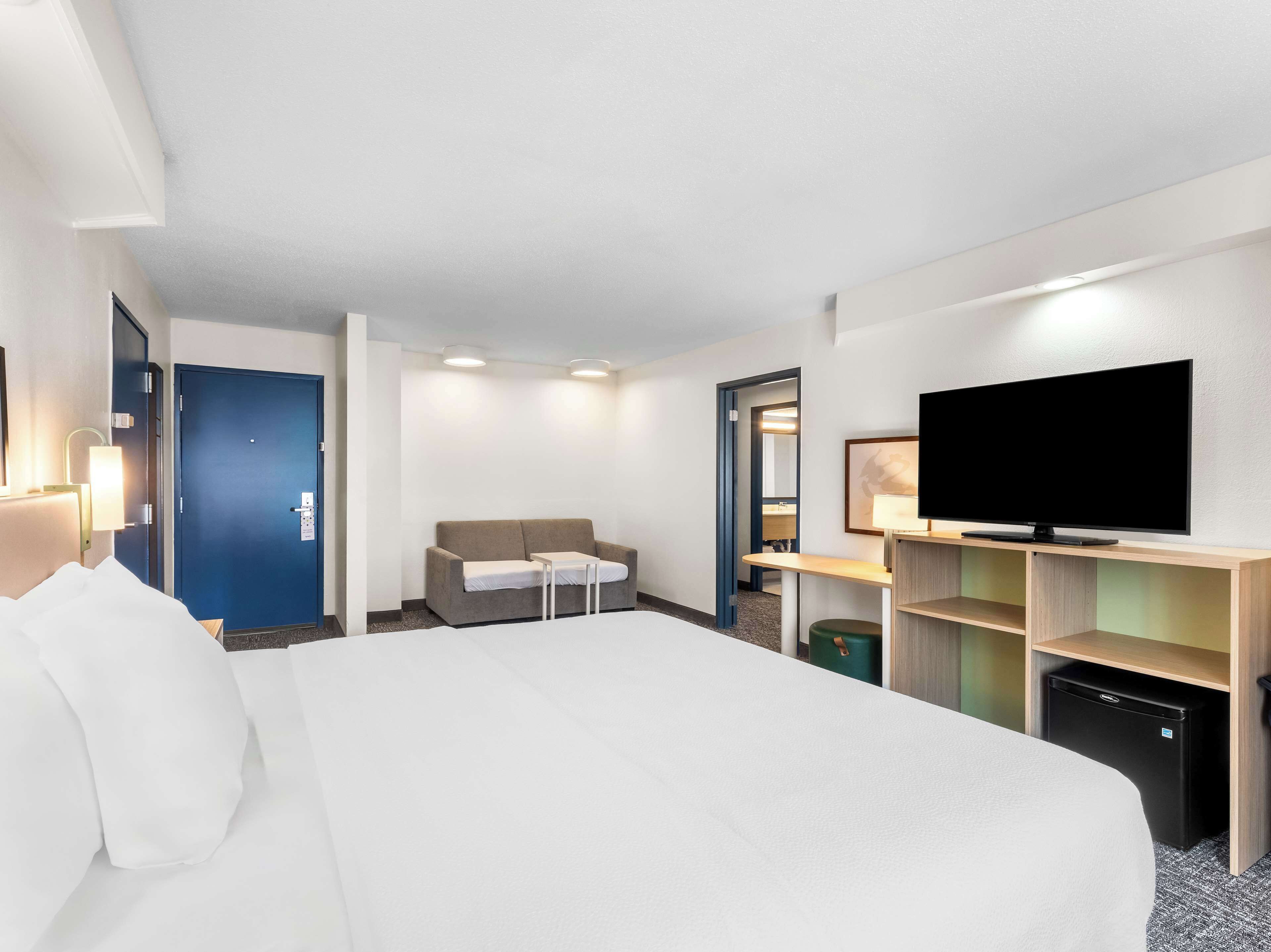 A hotel room with a large bed, a sofa, a TV, a desk, and a small fridge.