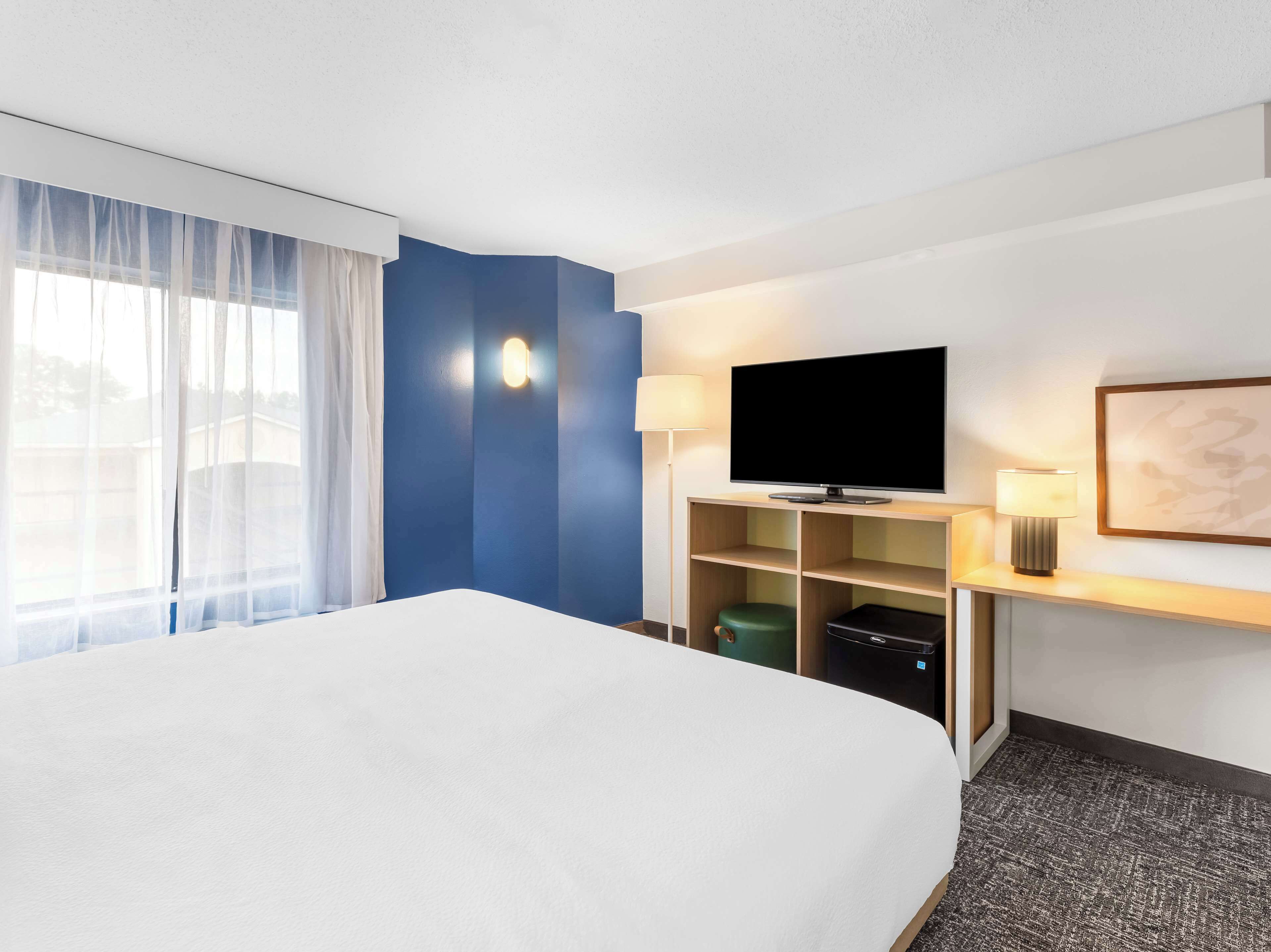 A hotel room with a large bed, a flat-screen TV, a desk with a lamp, and a wall-mounted shelf.