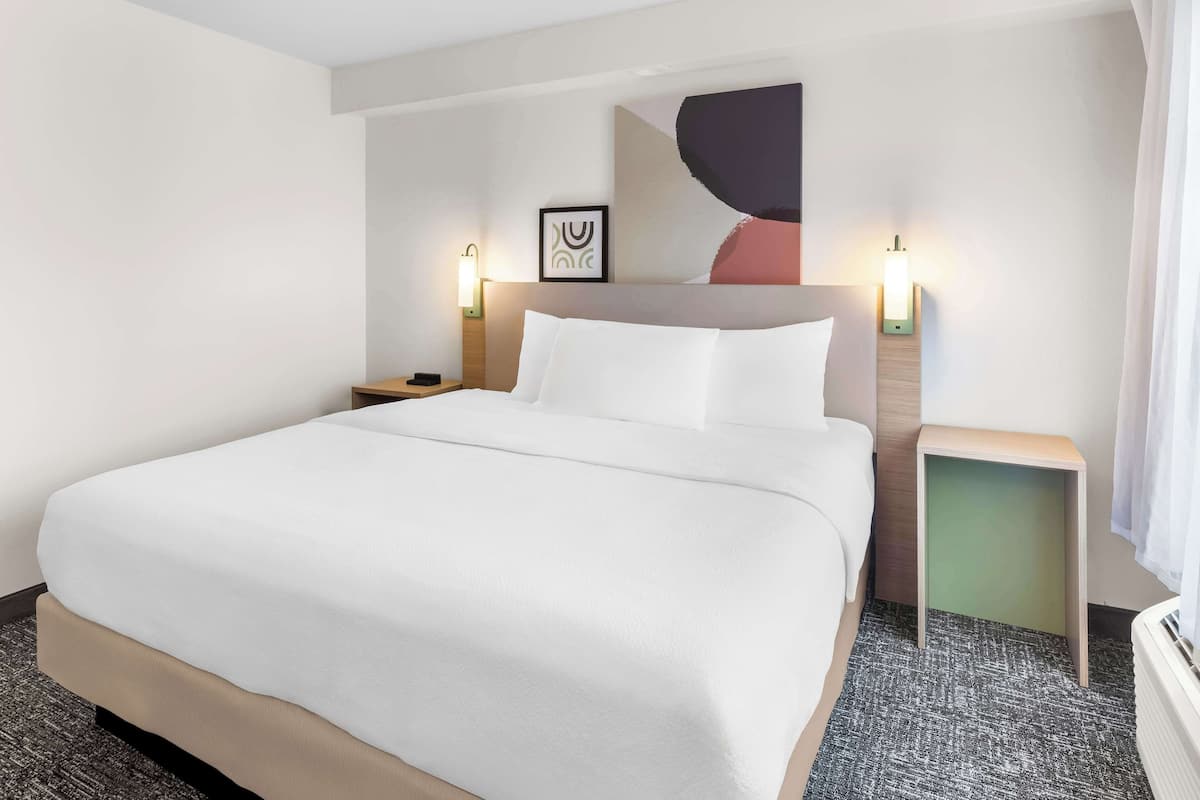 Una habitación de hotel con una cama grande, dos mesitas de noche, una obra de arte en la pared y un velador con lámpara.
