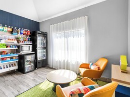 Una habitación moderna con una máquina expendedora llena de bocadillos, un refrigerador, una mesa redonda y dos sillas naranjas.