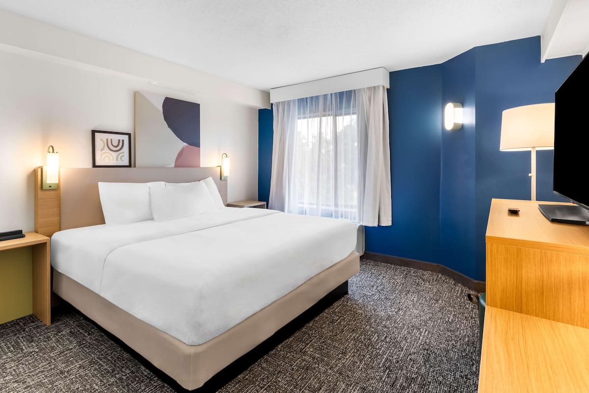 Una habitación de hotel con cama, mesitas de noche, una lámpara, televisión y ventana con cortinas.