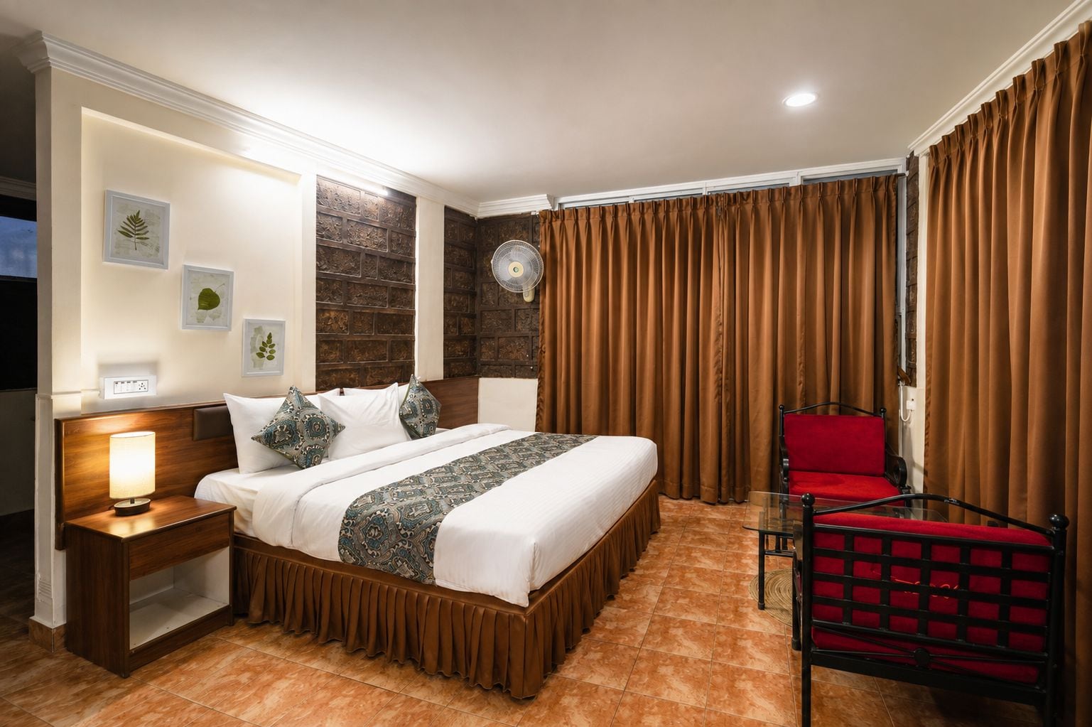 Una habitación de hotel con cama, mesita de noche, lámpara, una silla roja y cortinas.