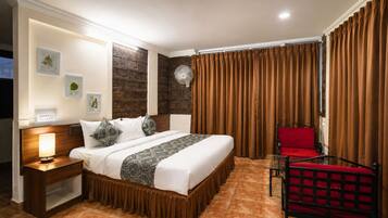Una habitación de hotel con cama, mesita de noche, lámpara, una silla roja y cortinas.