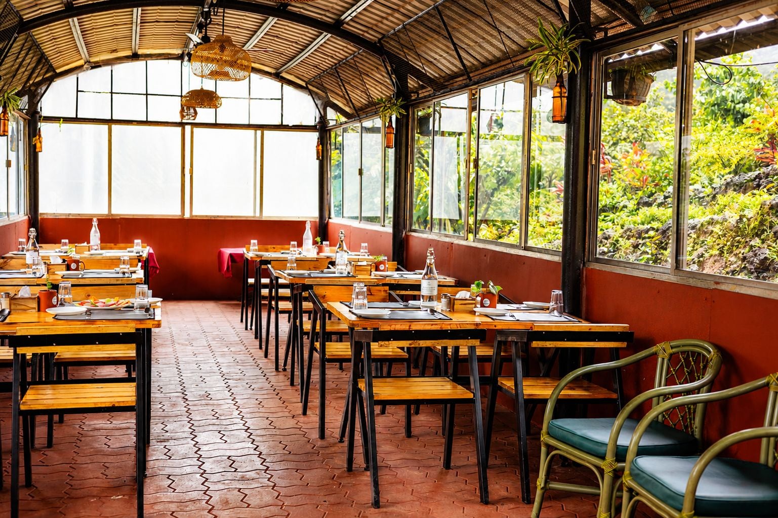 A restaurant with wooden tables and chairs, a red wall, and large windows overlooking greenery.